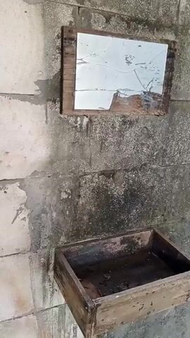 A cracked mirror is mounted on a concrete wall above a wooden box. The mirror is framed by wood and appears to be damaged. The wooden box is open and filled with dark liquid and debris. The concrete wall is weathered and stained. The scene appears to be outdoors.
