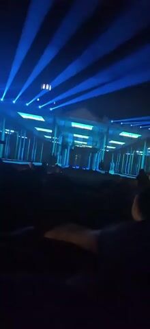 The video shows a nighttime outdoor event with a building facade illuminated by lights. Blue beams of light originate from the top left and project across the scene. The building in the background has a central section with a pediment and columns, and is decorated with vertical green and white lights along its facade. The building's exterior is also lit with a grid of yellow and white lights. There are people in the foreground, silhouetted against the light, appearing to be an audience watching the display. Some people are seated, while others are standing. The overall impression is of a light show or projection mapping event.