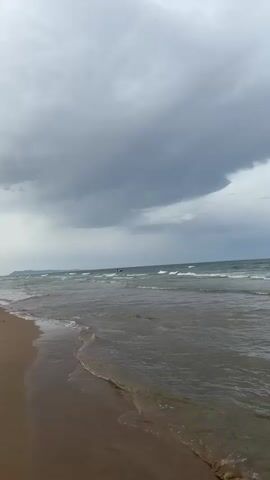 A view of a beach with gray clouds overhead and small waves lapping at the shore. In the distance, several kite surfers are visible on the water. The sand is wet from the tide.
