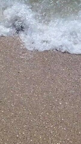 The video captures a serene beach scene at Calafell, Spain. The footage begins with a close-up of gentle waves lapping onto a sandy shore, showcasing the clear, shallow water and the glistening sand. As the camera pans out, the vast expanse of the blue sea stretches towards the horizon, under a clear sky suggesting a pleasant day.

In the distance, a large inflatable water park with slides and various structures is visible, indicating a recreational area. A few individuals can be seen enjoying the water park, and one person is swimming or playing in the sea closer to the shore. The gentle movement of the water and the bright sunlight create a peaceful and inviting atmosphere. There is no visible text in the media file. The overall impression is one of a relaxed summer day at the beach.
