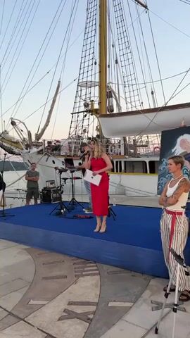 A woman in a red dress stands on a blue stage in front of a tall ship. She holds a microphone in her right hand and a clipboard in her left. She appears to be speaking to an audience. Behind her, a man wearing a baseball cap stands at a DJ setup with a laptop and audio equipment. Another woman with tattoos on her arms stands to the right of the stage, facing the ship. The sky is clear and the sun is setting.