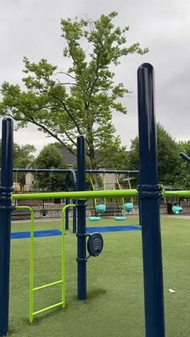 The video shows an empty children's playground with a swing set and other play equipment. The ground is covered in artificial turf, a mix of green and blue. Several trees with lush green foliage are visible in the background, along with some houses. The sky is overcast, suggesting a cloudy day. There are no people or animals visible in the footage.