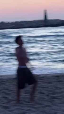 The video shows a young man on a sandy beach near the ocean at dusk. He is shirtless and wearing dark shorts, playing with a soccer ball. In the background, waves are breaking on the shore, and a pier with a lighthouse can be seen. The video also shows another young man briefly in the distance, also on the beach. The lighting suggests the sun has recently set.