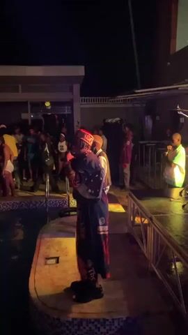A low-angle, medium shot captures a man wearing a long, dark robe with ornate designs and a red-and-gold hat. He stands on a platform next to a pool, holding a microphone and facing a crowd of people at an outdoor party at night. The lighting shifts between green and blue, illuminating the man and the surrounding area. A metal railing is to the right, and part of a building is visible in the background. Some partygoers are dancing.