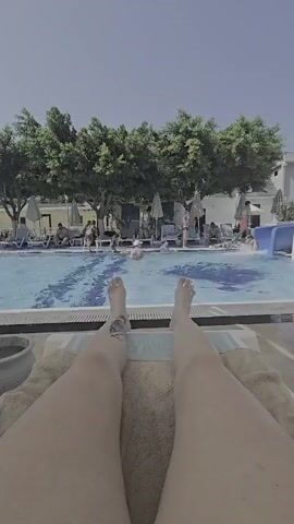The video is shot from a first-person perspective, looking down at the legs of a person lying on a lounge chair. Beyond the legs, a swimming pool is visible, with people swimming and lounging around it. Trees line the edge of the pool, and a building is visible in the background. The sky is clear and blue. The person's legs are bare, and they have a tattoo on their left calf. The scene appears to be a resort or hotel with a swimming pool. There is a water slide on the right side of the pool.