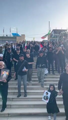 A crowd of men and one young girl are walking down a set of stone stairs. Many of the men are wearing black clothing and some are holding water bottles. One man is holding an Iranian flag. The young girl in the foreground is holding a framed picture of a man. The sky is visible in the background, and there are some buildings and flags in the distance.