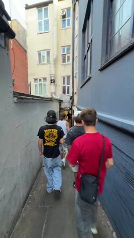 Four men walk single file down a narrow alleyway. The man in the front wears a black t-shirt and baggy blue jeans, and a dark bucket hat. The next man wears a red t-shirt, baggy gray pants, and a black crossbody bag.  The third man has on a dark short sleeved button up shirt and khaki pants. The last man has on a dark patterned shirt, white shorts and black sneakers with white socks.  The alley is between two buildings and has gray painted walls. There is some graffiti on the left wall. A metal pipe runs along the building on the left.