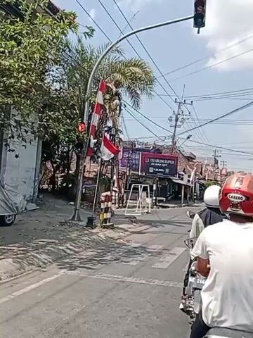 This video shows a street scene with a traffic light on a pole that is bent at an angle. The traffic light is red. There are power lines overhead. Two people on a motorcycle are visible in the foreground, stopped at what appears to be a crosswalk. In the background, there are shops and buildings. A flag with red and white stripes is attached to the bent traffic light pole.