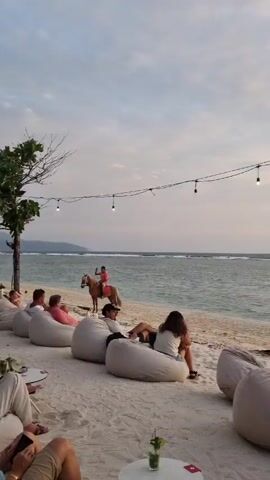 The video shows a beach scene at sunset. People are relaxing in beanbag chairs on the sand, with a horse and rider wading in the water nearby. As the sun dips lower, two women walk along the beach. One stops to take a photo, while the other seems to be talking on her phone. String lights hang above the beach. The sky is a mix of clouds and clear sky, with the setting sun painting the scene in warm colors.