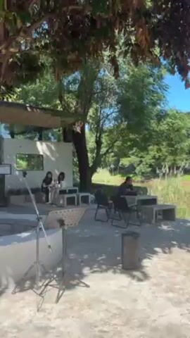 The camera pans across an outdoor cafe on a sunny day. Several groups of people are seated at tables made of concrete, chatting or looking at their phones. The tables are arranged in a partially shaded area beneath trees. A river runs along the edge of the cafe, with a wooded area on the opposite bank. The water in the river is muddy brown. Some people sit in chairs facing the river, while others are gathered at tables slightly further back.