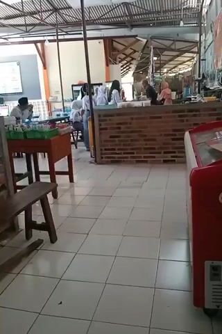 The video shows an indoor space that appears to be a food stall or a cafeteria. In the foreground, there are white tiled floors and a wooden bench. A red refrigerator is visible on the right side. In the middle ground, there are tables and chairs, some occupied by people. On the left, a person wearing a white shirt and a black cap is seated at a table with some food items in front of them. In the background, there is a counter made of brick and wood. Several people, including some wearing white uniforms, are standing behind the counter, appearing to be customers or staff. There are shelves and displays of food or drinks behind the counter. The overall lighting suggests it is daytime, and the space has a covered roof structure.
