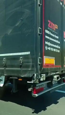 The video opens with a close-up of the side of a black truck with the partial logo "ZAUN" visible in white letters. The camera then pans to the right, showing the truck's wheel and the road. The scene shifts to a view of a grassy embankment and trees under a blue sky with some clouds. The final shots are from inside a car, looking out the side window and into the side mirror. The mirror reflects the road behind, showing the car traveling alongside another truck. The surrounding environment is a highway with a grassy verge.