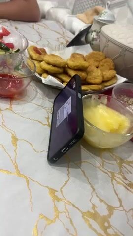The video shows a family enjoying a meal on a white tablecloth with gold marble-like accents. The table is laden with food, including a plate of chicken nuggets, a bowl of ketchup, a bowl of butter, and a salad of cucumbers and tomatoes. A cell phone stands upright amidst the food items, displaying a video game on its screen. A person uses a fork to dip a nugget into the ketchup. The rest of the group is out of focus and only partially visible. In the background are glimpses of decorative objects, including a teapot and a wicker container.