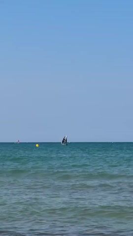 The video shows a wide shot of the ocean under a clear blue sky. There are small waves on the surface of the water. In the distance, there are a few windsurfers. One windsurfer in the center of the frame is more prominent, with a dark sail. To the left, another windsurfer with a red sail is visible. Further to the left, a yellow buoy is floating on the water. The horizon line is clearly defined, separating the blue of the sky from the green-blue of the sea. The overall scene conveys a sense of calm and leisure activity.