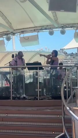 A live music performance takes place outdoors under a white canopy on a sunny day. The stage has a glass railing in front of it and is set up with musical instruments and equipment. On the left, a man wearing sunglasses and a pink collared shirt plays a guitar. Beside him, a keyboard is set up. To the right of the keyboard, a woman in a purple top and blue jeans is singing into a microphone and gesturing with her right hand. Behind them, other band members are visible, including a drummer. In the background, there are rows of tiered seating, suggesting an audience is present, and parts of a ship or large structure can be seen. The overall setting appears to be a deck area, possibly on a cruise ship, with a pool or deck chairs in the foreground.