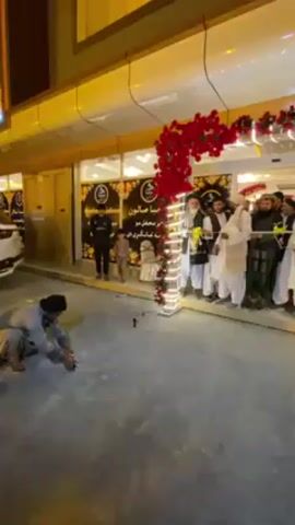 The video captures a nighttime scene during a celebratory event at the entrance of a building in Kandahar, Afghanistan. A group of men, primarily adults dressed in traditional Afghan attire such as shalwar kameez, turbans, and vests, are gathered at the main entrance. The doorway is elaborately decorated with red floral garlands and white string lights. A green ribbon is stretched across the entrance, held by several men, indicating a ribbon-cutting ceremony. An older man appears to be the central figure, poised to cut the ribbon, with a small tray containing scissors presented to him. In the foreground, a man is kneeling on the ground, lighting small cylindrical firecrackers, contributing to the festive atmosphere. A young boy is also visible among the attendees. To the right of the entrance, a white Lexus LX SUV is parked. A sign above the entrance displays text in Arabic or Persian script, including "سالون" (Salon) and "محافل" (mahāfil - events/gatherings), suggesting the establishment is a salon or a venue for gatherings. The overall mood is one of celebration and festivity.
