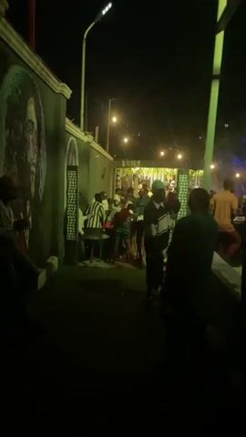 The video shows a crowded outdoor gathering at night, with the ambiance lit by strings of lights and colored stage lighting. People are dressed casually and appear to be socializing and dancing. In the foreground, individuals are seen moving and interacting, with some gathered around tables. The background reveals more people in a lively setting, possibly a bar or a party venue, with decorations and structures suggesting an established event space. The lighting shifts between green and blue hues, enhancing the festive atmosphere.