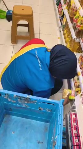 A person wearing a blue and yellow uniform with a dark blue hijab is crouching down in front of a store shelf, sorting items from a blue crate. The shelf is filled with various packaged goods, including bottles of cooking oil. To the left of the person is a tan stool. The floor is tiled. The person is picking up items from the crate and placing them on the shelf.