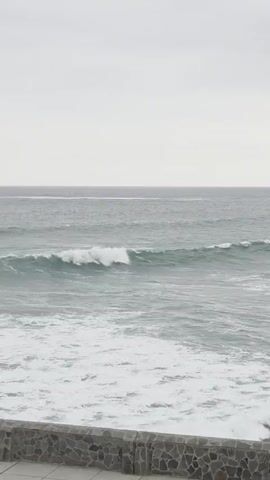 The video shows a wide shot of the ocean with gray skies. In the foreground, there is a stone wall. Waves are crashing on the shore, creating white foam. A person can be seen surfing in the distance. The overall atmosphere is calm and serene.