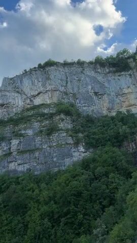 The video pans from a layered rock face with lush green vegetation to reveal a stunning mountain vista on a sunny day with a partly cloudy sky. A charred tree trunk stands in the foreground, partly obstructing the view of rolling green hills stretching into the distance.