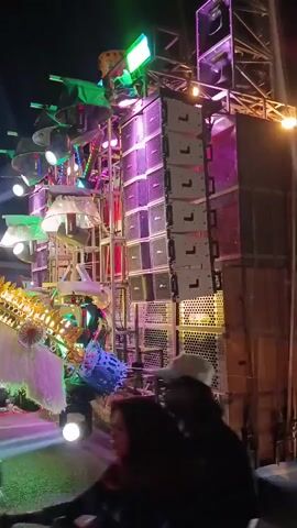 The video shows a large sound system with many speakers and colorful lights at night. Two people on a motorcycle are visible in the foreground, facing away from the camera. The sound system is elaborate, with speakers stacked high and numerous lights flashing and changing colors, creating a vibrant, energetic atmosphere. The overall setting appears to be an outdoor event or gathering.