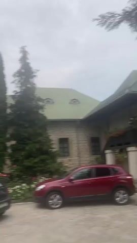 The video shows a panning shot of the exterior of a monastery or religious complex. In the foreground, there are cars parked on a paved area, and a rose garden with red, yellow, and white roses. The main building is made of stone with a green roof and arched windows. There are tall cypress trees and other evergreen trees surrounding the building. In one shot, Romanian flags are visible hanging from the building. Further in the video, a church with painted frescoes on its exterior walls and a tall tower is shown. The sky is overcast. There are no people visible in the video.