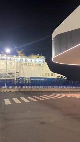A large white ferry with blue accents is docked at a terminal at night. The ferry is illuminated by lights from within and from a bright spotlight on the left side. The name "BUENAVISTA EXPRESS" is visible on the side of the ferry. A modern building with a curved, illuminated facade is to the right of the ferry. In the foreground, there is a dark asphalt surface with white pedestrian crosswalk markings. Behind the crosswalk, there is a blue fence with white lettering that appears to say "FRED OLSEN". A dark-colored car is parked behind the fence. The sky is dark and clear.