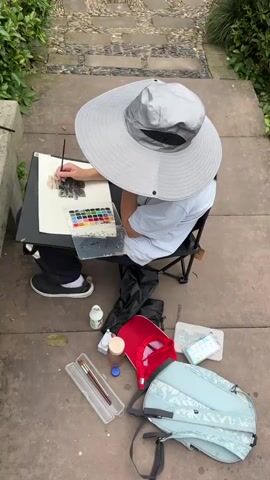 The video shows a person painting a waterfall scene. The person is sitting on a small folding chair, wearing a large brimmed hat, a white t-shirt, and dark pants with black and white shoes. They are using a watercolor palette and a brush to paint on a piece of paper attached to a board. Around the person are various art supplies, including paint bottles, brush cases, and a backpack. The camera then shifts to show the waterfall. The waterfall is made of stone and covered in green plants. The water cascades down the rocks into a small pool below. The sound of the waterfall can be heard throughout the video.