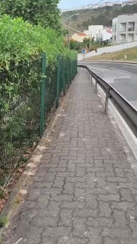 The video shows a pedestrian view of a paved sidewalk next to a green metal fence with overgrown bushes. The sidewalk is made of interlocking concrete pavers and has some debris on the edge. To the right of the sidewalk is a road with a metal guardrail separating it from the sidewalk. Cars are visible driving on the road, and buildings are visible in the background on a hillside. The sky appears overcast. The camera is moving forward along the sidewalk.