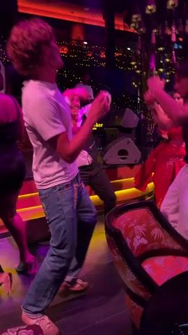 The video shows a group of people dancing in a dimly lit club. The lighting is predominantly pink and red, with some gold accents. In the foreground, a man with curly brown hair wearing a white t-shirt and blue jeans is dancing. To his left, a woman in a black dress is also dancing. Behind the man in white, a bald man wearing a vest is dancing and smiling. To the right of the man in white, a woman in a red sequined dress is dancing. In the background, more people are visible dancing, and there are decorative lamps hanging from the ceiling. The overall atmosphere appears to be lively and festive.