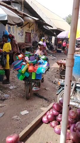 The video captures a daytime scene at an outdoor market in Akwa Etiti, Nigeria. In the foreground, a wooden stall displays a large pile of red onions. A woman, wearing a white cap and a dark top, is pushing a wheelbarrow laden with various colorful plastic household items, including bowls, cups, and baskets. She moves along an unpaved dirt path, past simple market structures with corrugated metal roofs.

In the background, other people are visible, including a woman in a yellow top on the left and another in a pink top further down the path. Various market elements such as yellow jerry cans, a blue barrel, and a motorbike can also be seen. The bright sunlight suggests it's a clear day, and the overall atmosphere is that of a busy, functional market with vendors transporting and selling their goods. Text is visible on some signs on the buildings in the background, but it is too blurry to decipher.