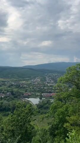 A wide-angle, elevated view showcases a verdant valley landscape under a cloudy sky. The camera pans slowly from left to right, revealing a town or village nestled in the valley, surrounded by lush green trees and rolling hills. Numerous buildings with various roof colors are visible, interconnected by roads that wind through the settlement. A body of water, possibly a lake or a wide river, is prominent in the mid-ground, reflecting the muted light from the overcast sky. Farmland or open green spaces are also interspersed within the village and the surrounding landscape. Dense green foliage from trees fills the foreground, partially framing the panoramic vista. In the distance, several layers of mountains or large hills extend towards the horizon, gradually fading into the atmospheric haze. The overall impression is one of a peaceful, expansive natural setting with a small community integrated into it. No people or visible text are present. The weather appears to be overcast but bright, indicative of daytime.