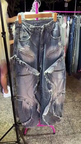 The video shows a close-up of a pair of distressed, wide-leg denim jeans hanging on a clothing rack. The jeans are a light gray-blue wash with extensive rips, tears, and frayed edges, particularly around the thighs and lower legs. There are zippered pockets on the front of the thighs. The background shows a row of other clothing items on hangers and a blurry view of an outdoor market at night with lights. A person's hands are visible in the foreground, interacting with the clothing. The audio includes indistinct chatter and possibly music, and a voice says "push boom, all for 20".