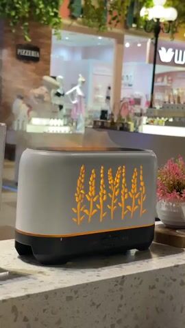 The video shows a close-up of a white and black humidifier with an orange wheat stalk design on its side. Mist is emitting from the top of the humidifier. The humidifier is placed on a grey speckled countertop. In the background, there is a blurred scene that appears to be inside a mall or a retail space. There are storefronts visible, one with a sign that says "PIZZERIA". Mannequins and merchandise are also visible in the background, as well as a small potted plant with pink flowers. A number tag with "890" is visible in the foreground, next to the humidifier. The lighting appears to be artificial, creating a warm ambiance.