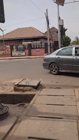 The video shows a street scene, seemingly in an urban environment. The camera begins pointed towards a street with a manhole cover in the foreground, and then pans left, revealing a small roadside business operating under an umbrella, supported by wooden structures and a box. A person wearing what appears to be a security uniform is sitting on a bench, seemingly looking at their phone. The surrounding area has utility poles, and some foliage, including pink flowers and a small tree.