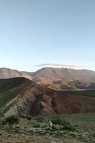 The video shows a view of a lake surrounded by hills and mountains under a clear sky. The foreground features a grassy hill with some bushes. The lake stretches into the distance, its calm surface reflecting the sky. Mountains rise in the background, some shrouded in a soft haze. The lighting suggests it's either early morning or late afternoon. There's a child audible, exclaiming "wahh."