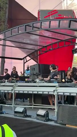 A group of people are on a stage behind DJ equipment. A woman wearing headphones is at the center, facing a set of turntables and a mixer. She is wearing sunglasses and a black t-shirt and skirt. To her left, a man in a black t-shirt and sunglasses is also at a DJ station. To her right, several people are watching and some are raising their hands. Behind the DJ equipment, a large red structure is visible. The stage is covered by a transparent roof with metal beams. There are speakers on the stage and in front of it. The overall impression is of an outdoor music event.