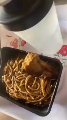 A person's perspective video shows a take-out container of spaghetti and chicken sitting on a table. A large water bottle is situated above the take-out container. The spaghetti has a red sauce. The chicken appears to be a browned chicken thigh. The container is black plastic. The table cover is white and decorated with red flowers.