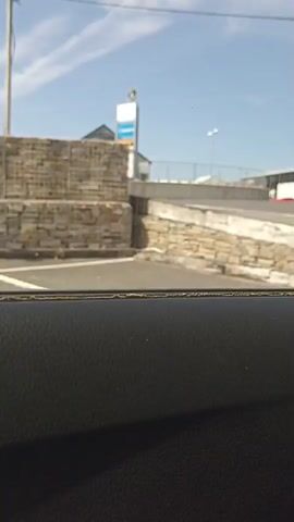 The video begins by showing a black textured surface, possibly the interior of a car. The camera then pans right to reveal an outdoor scene. In the background, there is a light blue sky with some wispy clouds. There are several buildings visible, including one with a light-colored, horizontally paneled facade. A road or pathway curves upwards towards this building. A metal gate with yellow and white reflective strips is partially obstructing the view of the pathway. This gate appears to be a barrier, possibly for a parking lot or an entrance. There are also some stone walls and a lamppost visible. The sun is shining, casting shadows, indicating it is daytime. The overall scene suggests a road or an access point leading to some kind of facility.