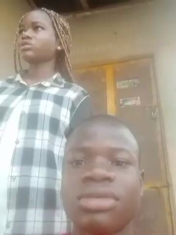 A video shows two young people, likely a boy and a girl, standing together outdoors. The boy is positioned in the foreground, while the girl stands slightly behind him. He is looking directly at the camera with a smile, while she appears to be looking off to the side and holding a mobile phone in her left hand. The girl has her hair styled in braids and is wearing a green and gray plaid shirt. Behind them, there's a light-colored wall with some markings or writing on it and a yellow door with some signs attached. The lighting suggests it's daytime, but the setting and exact location are unclear. The boy is speaking in the video.