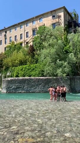 The video shows a group of people enjoying a day at a river spot. A group of people are standing in a shallow part of the river, while others are swimming in a deeper section. Some people are standing on a low stone wall, jumping into the water. The river is a clear turquoise color and flows over a rocky bed. In the background, there is a building and a hillside covered with trees. The sky is clear and blue.