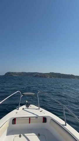 This is a first-person perspective video from the bow of a white motorboat traveling on the open ocean. The water is a deep blue with moderate waves and ripples. In the distance, a rugged, tree-covered coastline with buildings is visible under a clear, bright blue sky. The boat's white deck, cushioned seating with red and white accents, and chrome railings are in focus in the foreground. The camera is static, pointing forward, and the boat appears to be moving steadily. There are no people visible in the shot.