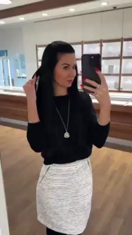 A woman is taking a selfie in a mirror in a store. She is wearing a black long-sleeved shirt, a white and gray striped skirt, a long silver necklace with a pendant, and two gold bracelets. She has long black hair and red nail polish. She is looking at her phone, which is a dark color. The store has wooden floors and cabinets with jewelry on display behind her. There is a security camera in the upper right corner of the frame.