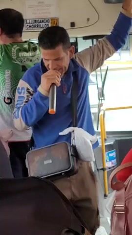 A man wearing a blue and beige long-sleeved shirt and brown pants is singing into a microphone on a bus. He has a black bag slung across his body. The bus is crowded with other passengers. One passenger in the background wears a green shirt with a logo.
