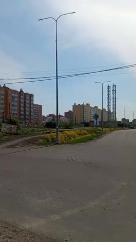 The video shows a street view in a residential area on a clear day. The camera pans from an intersection with apartment buildings in the background to a road leading into a field of yellow flowers. There is a dirt shoulder alongside the road, with utility poles and wires visible along the roadside. There is little traffic on the road. The sky is blue with some clouds.