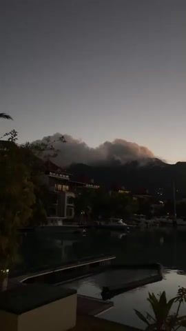 A vertical video shows a serene harbor scene at dusk. The sky transitions from a deep twilight gray to hues of orange and yellow as the sun sets behind a mountainous horizon. Clouds are clustered around the peaks of the mountains, catching the last rays of sunlight, appearing illuminated in pink and orange. Below the mountains, a row of buildings with red roofs is visible, nestled amongst lush green trees. The buildings are dimly lit, suggesting it's nighttime or late evening. In the foreground, a calm body of water reflects the sky and the surrounding landscape. Several docked boats, including yachts and smaller vessels, are visible on the water, their outlines dark against the illuminated water. Wooden docks extend into the water. A small potted plant with large leaves is visible in the bottom right corner of the frame, adding a touch of nature to the foreground. The overall atmosphere is peaceful and tranquil.