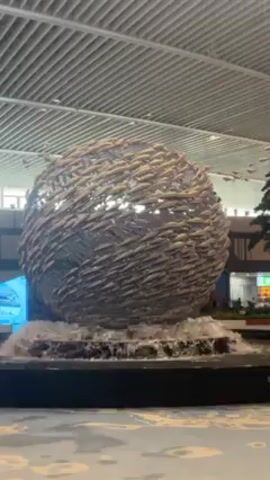 The video shows a large, spherical sculpture in an indoor setting, most likely an airport or shopping mall, given the ceiling architecture. The sculpture appears to be made of numerous smaller, silver-colored fish-shaped objects arranged in a circular pattern. The sculpture is situated in a fountain, with water flowing around its base. The lighting within the sculpture changes from white to purple. There are hints of other architectural elements in the background, including a blue light panel and a plant.