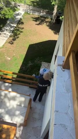 Two people are visible from a high angle, standing on a tiled outdoor area next to a white wall and a wooden railing. One person, wearing a black jacket with "CALVIN KLEIN JEANS" written on the back, is facing the wall and appears to be interacting with the wall. The other person, wearing a blue shirt and jeans, is bent over near the first person, also facing the wall. A wooden staircase is to the left of the scene, with a white couch visible on the steps. Beyond the tiled area is a grassy lawn with trees and shadows. The lighting suggests it is daytime.