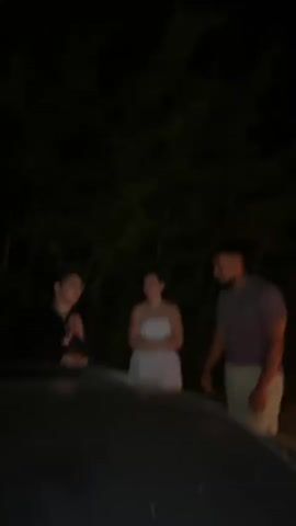 A dark outdoor scene features three people standing near a car. On the left, a person with glasses in a dark shirt smiles and holds a silver spray can. In the center, a woman in a white strapless top looks towards the person on the left. On the right, a man in a purple shirt and light-colored shorts faces the other two, gesturing with his right hand as if speaking. The background is filled with dark trees, and a car's dark hood is visible in the foreground, obscuring the bottom of the frame. The lighting is dim, suggesting it is nighttime.