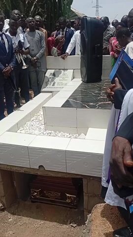 The video captures an outdoor funeral or burial service in Ladura Foron, Nigeria, under bright, sunny conditions. A diverse group of people, predominantly men, are gathered around a white-tiled grave structure, which features several levels and white gravel.

In the foreground, a dark, ornate casket, possibly wooden with gold-colored details, is visible within a partially dug grave pit beneath the tiled structure. A black portable speaker rests on the grave.

A clergyman, dressed in a white robe and purple stole, is officiating the service, speaking into a microphone while holding a book or binder. Attendees stand respectfully, many in formal or semi-formal attire, including some in white shirts and black trousers who might be security personnel. One man holds a trumpet, suggesting a musical element to the ceremony. Another individual in a black hoodie is seen operating a DSLR camera, documenting the event. The overall mood appears somber and respectful.

Visible text on the gravestone reads: "In loving memory of our beloved father," followed by "COFIA [partially obscured] ADEWANG" and dates "1954 - 2024," indicating the deceased was "Aged 70 Years." The background shows simple structures with corrugated iron roofs and some sparse vegetation, consistent with a semi-urban or rural setting.